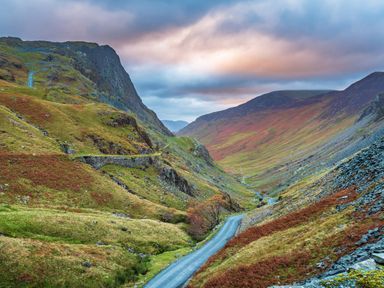 A winding road through a valley with grassy hills and rocky slopes under a cloudy sky at Rayside Ullswater