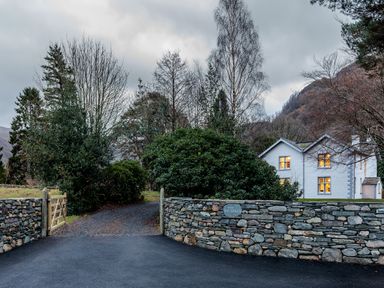 A house with lit windows behind a stone wall and a gate with trees and hills at The Vicarage near Rosthwaite