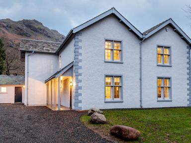 A white two-story house with lit windows and a gravel driveway near a grassy area with rocks at The Vicarage near Rosthwaite