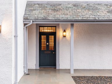 A dark green front door with glass panels under a small covered porch with a lit wall lantern at The Vicarage near Rosthwaite