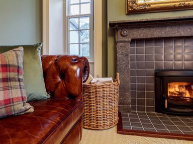 A brown leather sofa with cushions next to a wicker basket with firewood by a tiled fireplace at The Vicarage near Rosthwaite
