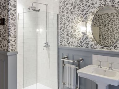 A bathroom with a tiled shower, floral wallpaper, a round mirror, a wall light, a radiator, and a pedestal sink at The Vicarage near Rosthwaite