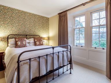 A bedroom with a metal bed frame, patterned wallpaper, pillows, bedside table with lamp, and large windows with curtains at The Vicarage near Rosthwaite