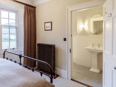 A bedroom with a metal bed frame radiator and curtain next to a window and an adjoining bathroom with a pedestal sink and round mirror at The Vicarage near Rosthwaite