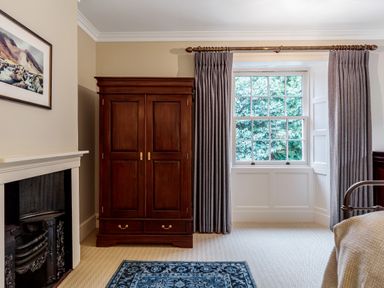 A bedroom with a wooden wardrobe next to a window with curtains and a fireplace with a framed picture above at The Vicarage near Rosthwaite