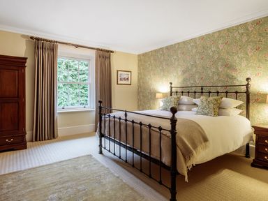 A bedroom with a metal bed frame pillows floral wallpaper wooden wardrobe and window with curtains at The Vicarage near Rosthwaite