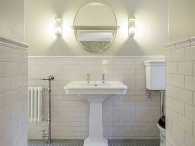 A bathroom with a pedestal sink a round mirror two wall lights a radiator and a toilet at The Vicarage near Rosthwaite