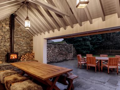 A covered outdoor dining area with a wooden table and benches with fur throws and a wood stove next to a stone wall at The Vicarage near Rosthwaite
