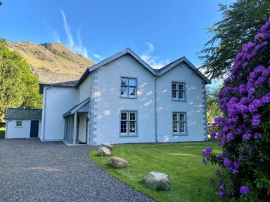 A white house with blue trim surrounded by grass and rocks with purple flowers on the side at The Vicarage near Rosthwaite