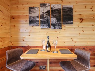 A small dining area with a wooden table two chairs a bottle and two glasses with a deer picture on the wall at Stag Lodge Pod in Llanfair Caereinion