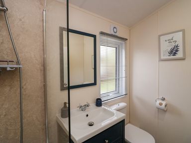 A bathroom with a sink, mirror, toilet, window with blinds, shower, and wall art at Lakeland Dream @ South Lakeland Leisure Lodge in Warton