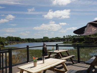 A wooden deck with picnic tables and flower pots overlooking a lake with cabins and trees at Lakeland Dream @ South Lakeland Leisure Lodge in Warton
