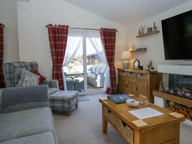 A living room with a plaid armchair and ottoman a wooden coffee table a wooden sideboard with lamps and decor and a wall-mounted TV at Lakeland Dream @ South Lakeland Leisure Lodge in Warton