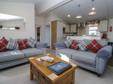A living room with two grey sofas with plaid cushions and a wooden coffee table in front of a kitchen at Lakeland Dream @ South Lakeland Leisure Lodge in Warton