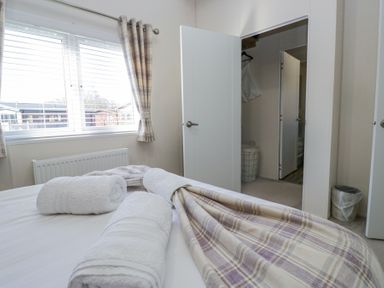 A bedroom with rolled white towels on a bed a window with plaid curtains and two open white doors showing a laundry basket and trash bin at Lakeland Dream @ South Lakeland Leisure Lodge in Warton