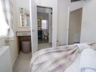 A bedroom with a bed, towel, plaid curtains, vanity table with stool, and open doors leading to a bathroom and another room at Lakeland Dream @ South Lakeland Leisure Lodge in Warton