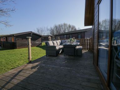 An outdoor deck with wicker furniture and a dining table next to wooden cabins at Lakeland Dream @ South Lakeland Leisure Lodge in Warton