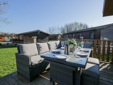 An outdoor dining area with a glass table set with plates and glasses on a wooden deck at Lakeland Dream @ South Lakeland Leisure Lodge in Warton