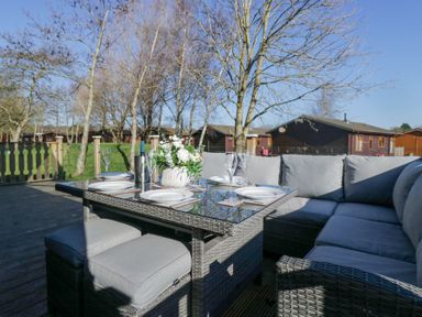 An outdoor patio with a glass top table set with plates and glasses surrounded by a cushioned sectional sofa and stools at Lakeland Dream @ South Lakeland Leisure Lodge in Warton
