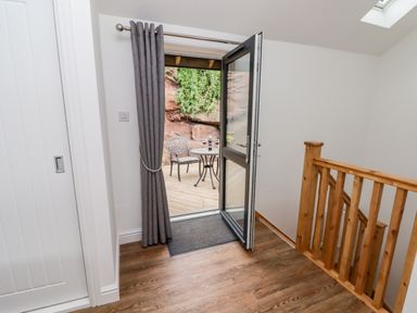 A hallway with wooden floor and railing leading to an open door with gray curtains and outdoor seating at The Old Mill Bake House in Goodrich