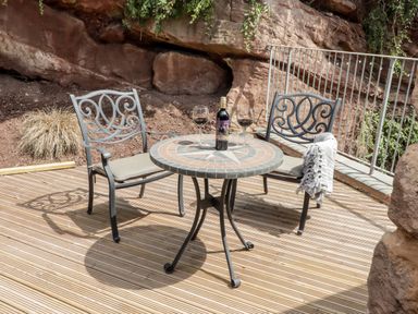 An outdoor patio with a round mosaic table and two metal chairs with cushions and a bottle of wine with two glasses at The Old Mill Bake House in Goodrich
