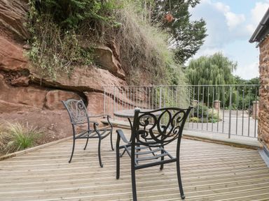 An outdoor wooden deck with two metal chairs and a round table near a rock wall and metal railing at The Old Mill Bake House in Goodrich