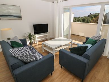 A living room with two blue sofas a white coffee table a television on a stand a floor lamp a plant and sliding glass doors leading to a balcony at 3 Lon Y Dryw in Trearddur Bay