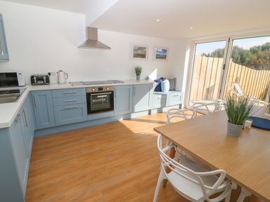 A kitchen with blue cabinets a wooden floor a dining table with white chairs and sliding glass doors at 3 Lon Y Dryw in Trearddur Bay