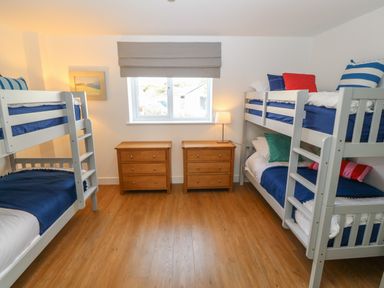 A bedroom with two white bunk beds and two wooden dressers with a lamp near a window at 3 Lon Y Dryw in Trearddur Bay