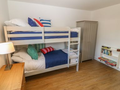 A bedroom with bunk beds a wooden dresser a floor lamp a wardrobe and a bookshelf with board games and books at 3 Lon Y Dryw in Trearddur Bay