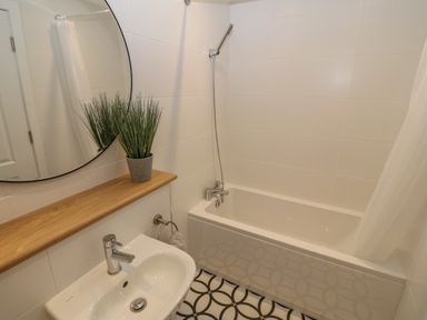 A bathroom with a bathtub shower combination a sink a round mirror and a potted plant on a wooden shelf at 3 Lon Y Dryw in Trearddur Bay
