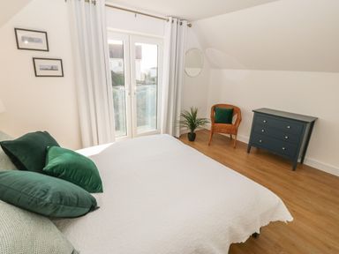 A bedroom with a bed and green pillows near glass doors with curtains a wicker chair a small plant and a dark blue chest of drawers at 3 Lon Y Dryw in Trearddur Bay