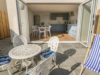 An outdoor patio with metal table and chairs with cushions and a blue striped deck chair next to a kitchen and dining area at 3 Lon Y Dryw in Trearddur Bay