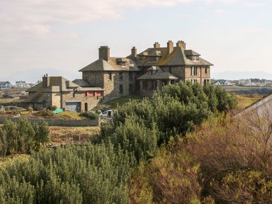 A large stone house with multiple chimneys surrounded by greenery and other distant houses at 3 Lon Y Dryw in Trearddur Bay