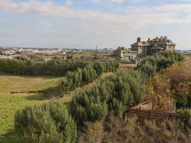 A large stone house surrounded by bushes and grassy fields with more houses in the distance at 3 Lon Y Dryw in Trearddur Bay