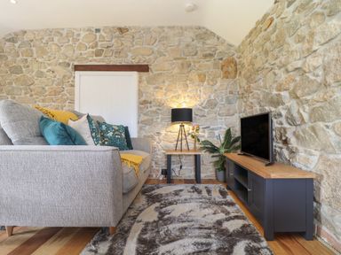 A living room with a gray sofa and decorative pillows a black lamp on a small wooden table a television on a wooden cabinet and stone walls at The Hayloft in Helston