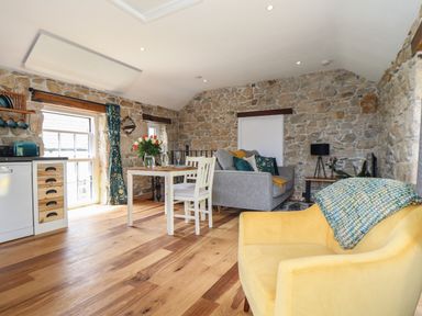 An open room with a yellow armchair a gray sofa a dining table and kitchen area with stone walls at The Hayloft in Helston