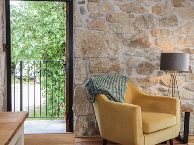A stone wall interior with a yellow armchair draped with a blanket next to a floor lamp and an open door showing a green tree outside at The Hayloft in Helston