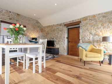 A room with a dining table and chairs a yellow armchair a small television on a stand and stone walls at The Hayloft in Helston