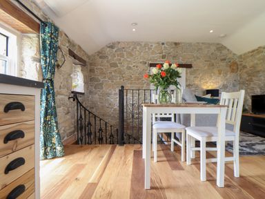 A dining area with a white table and chairs on a wooden floor next to a stone wall and staircase at The Hayloft in Helston