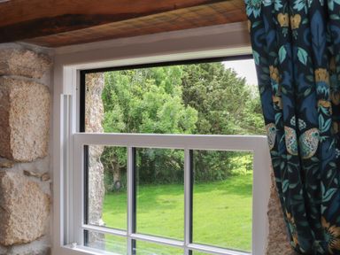 A window with white frame partially open showing green trees and grass outside at The Hayloft in Helston
