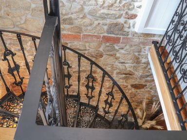 A black metal spiral staircase next to a stone wall with a wooden floor at the top and dried plants at The Hayloft in Helston