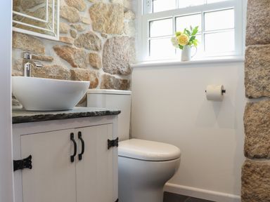 A bathroom with a stone wall a white cabinet with sink and a toilet below a window with flowers at The Hayloft in Helston
