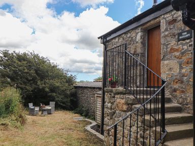 An outdoor seating area with wicker chairs and a table near stone stairs leading to a wooden door at The Hayloft in Helston