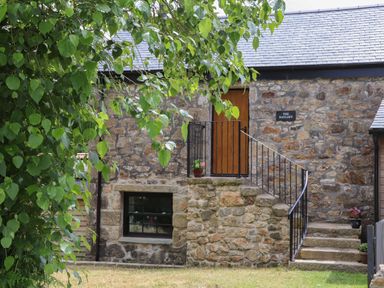 An exterior stone wall of a building with a wooden door accessed by stone stairs and a window beneath surrounded by green leaves at The Hayloft in Helston