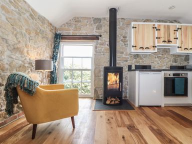 A room with stone walls a yellow armchair a floor lamp a stove with fire wooden cabinets and a window at The Hayloft in Helston