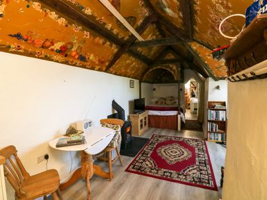 A room with a bed in the corner a wooden table with chairs a wood stove a bookshelf and a red patterned rug at Toad Hall Cottage in Hutcherleigh near Blackawton