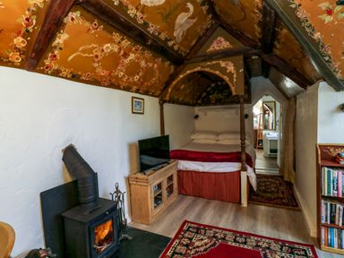 A bedroom with a bed under a decorated wooden ceiling a television on a cabinet and a wood stove in the corner at Toad Hall Cottage in Hutcherleigh near Blackawton