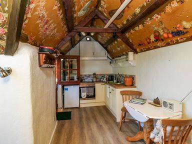 A small kitchen with a wooden floor a painted ceiling wooden beams a table with two chairs and kitchen cabinets at Toad Hall Cottage in Hutcherleigh near Blackawton