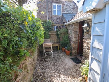 A narrow outdoor patio with gravel ground a small table and two chairs potted plants and bushes beside a stone house at Toad Hall Cottage Hutcherleigh near Blackawton